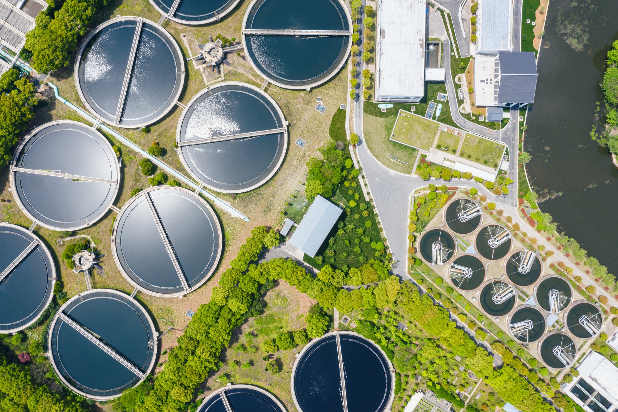 Aerial view of a wastewater treatment facility showing multiple circular clarifiers, aeration basins, and interconnected process infrastructure surrounded by landscaped grounds.