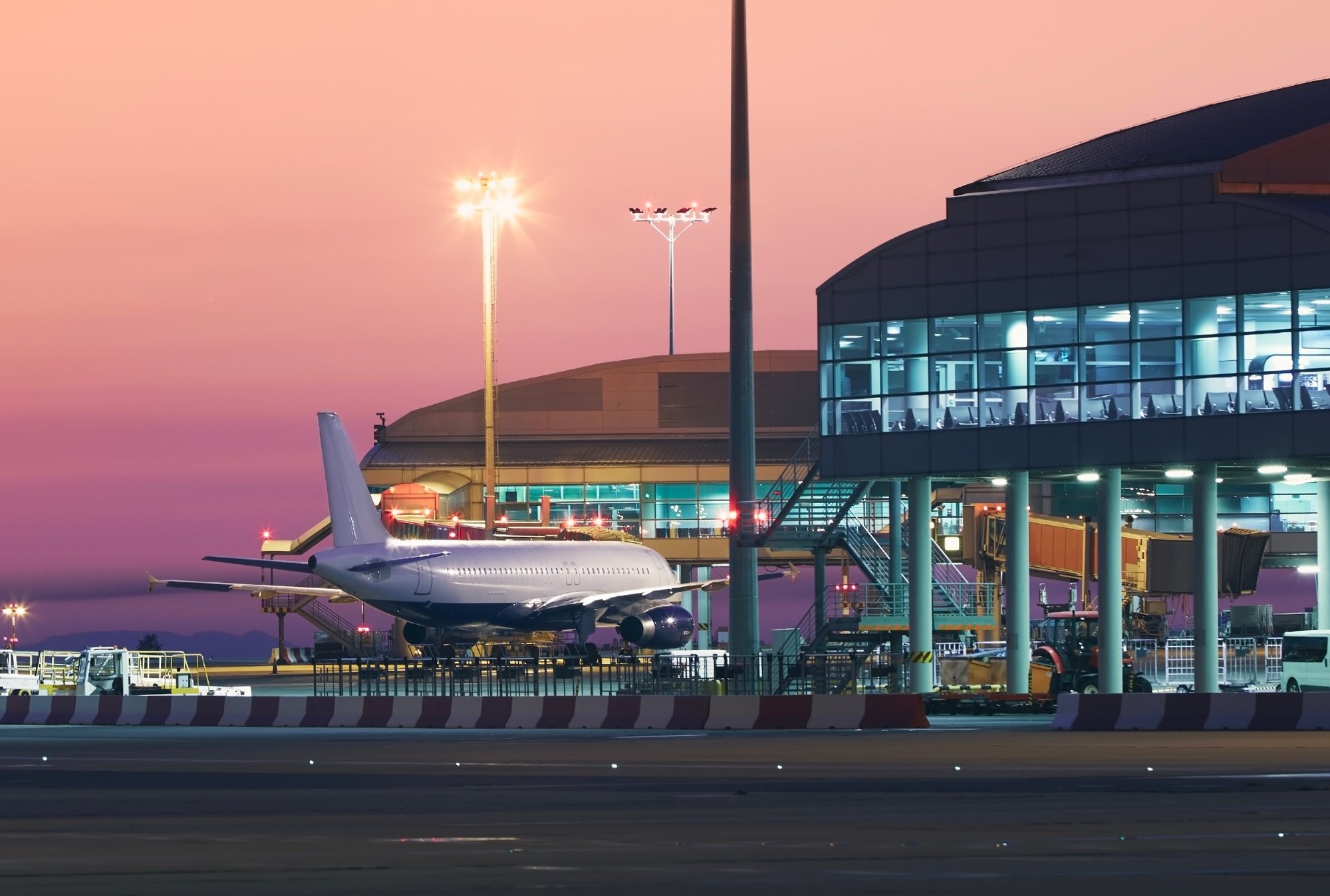 Commercial aircraft parked at an airport gate during dusk, with terminal buildings illuminated, ground support equipment visible, and airfield lighting highlighting ongoing operations.