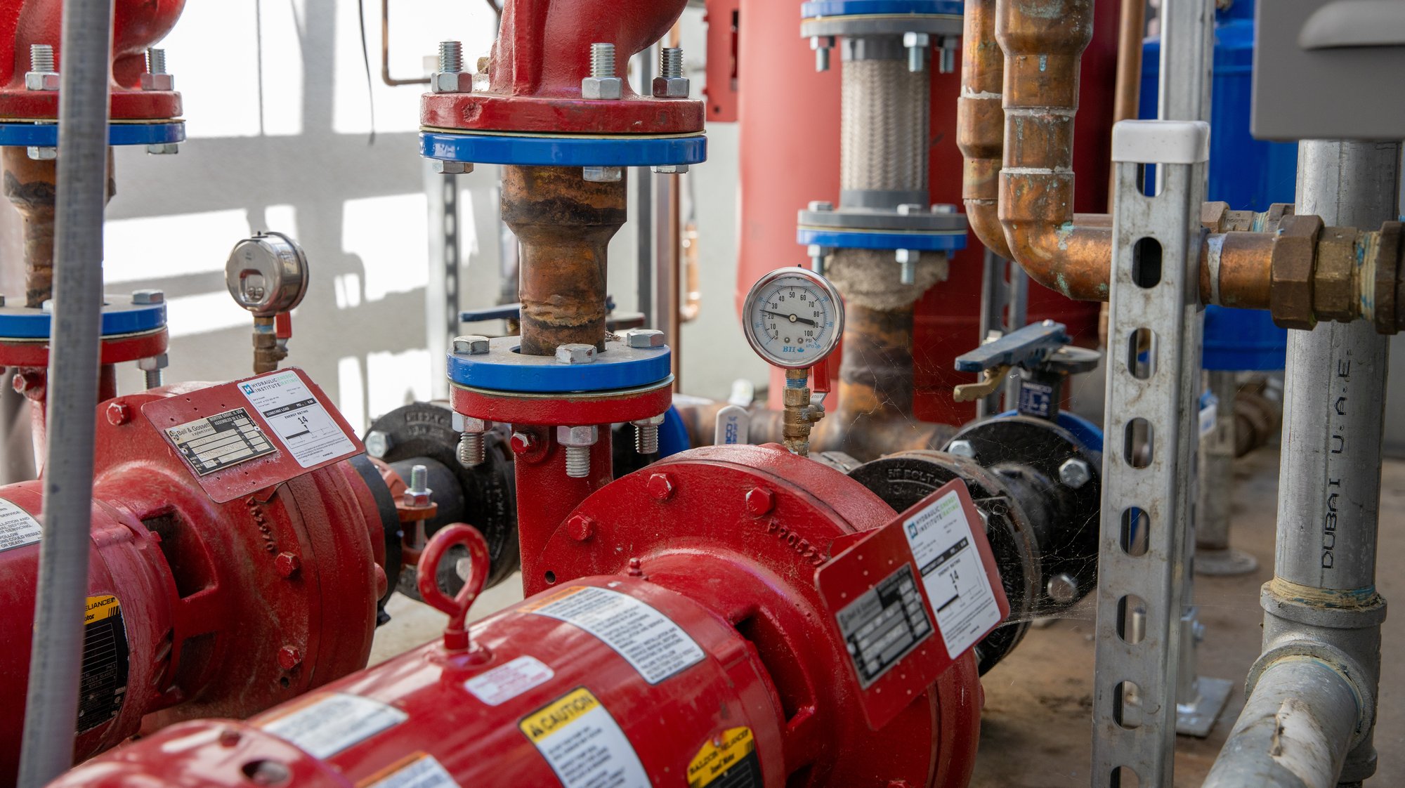 A mechanical room with red fire suppression pumps connected to copper and brass piping, multiple pressure gauges, isolation valves, and a vertical gauge panel mounted on the wall.