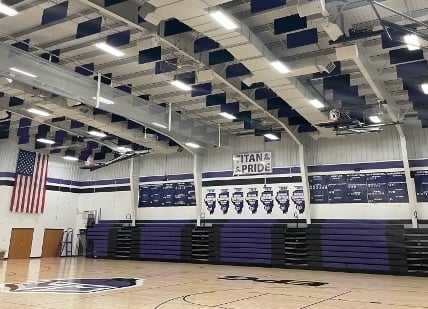 Interior of a school gymnasium with basketball court, bleachers, acoustic ceiling panels, and banners on the walls.
