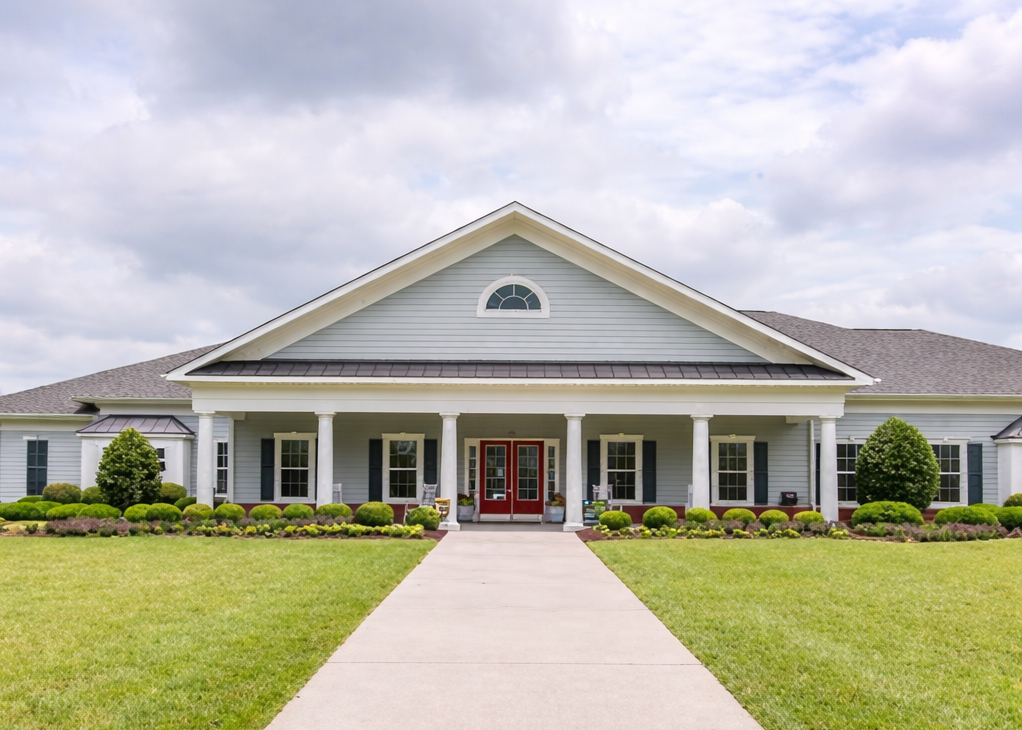 Exterior view of a military housing community building with a covered entrance, white columns, landscaped grounds, and a central walkway leading to red double doors.