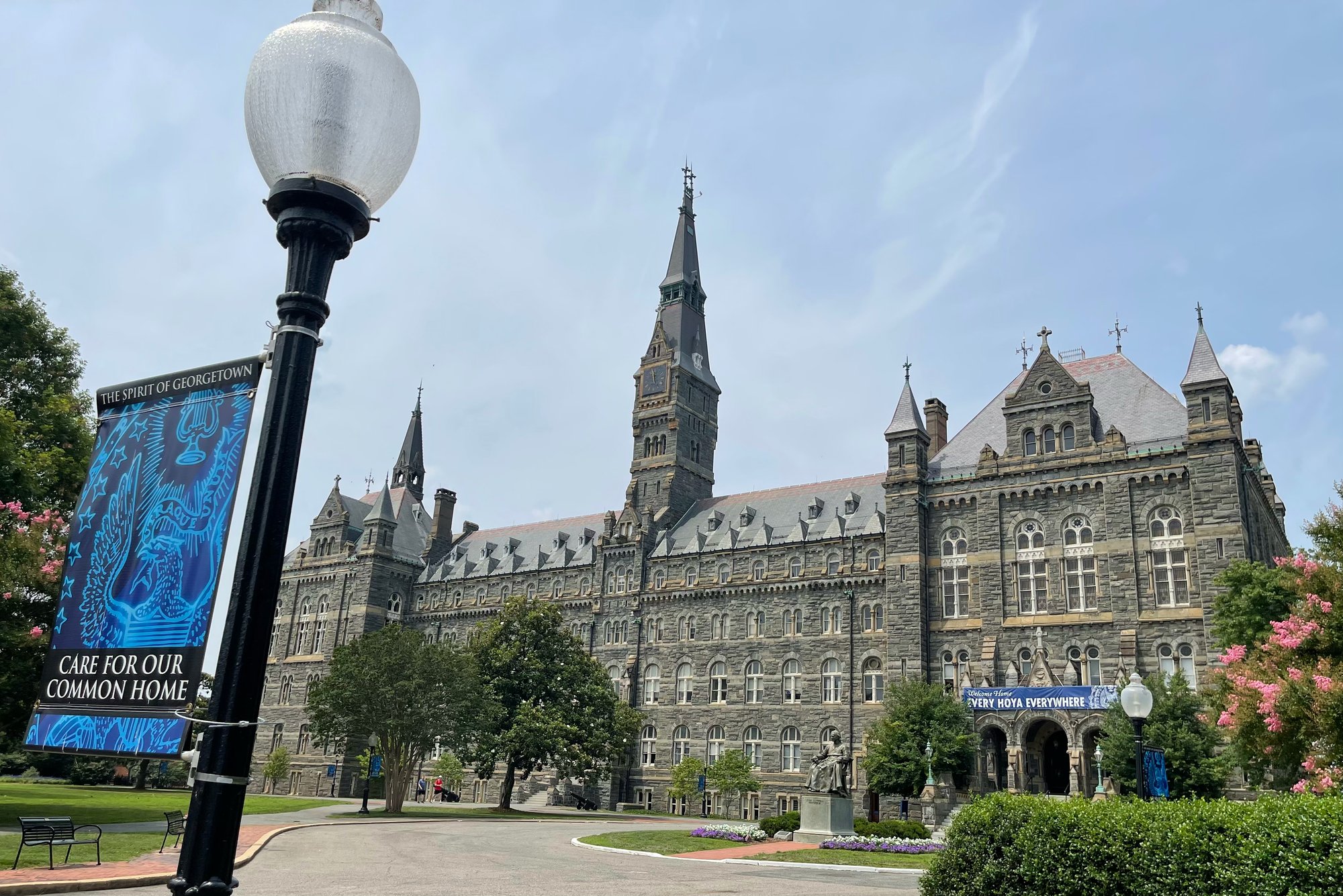 Historic stone academic building with a central clock tower at Georgetown University, viewed from a landscaped campus walkway with lamppost banner and surrounding greenery.