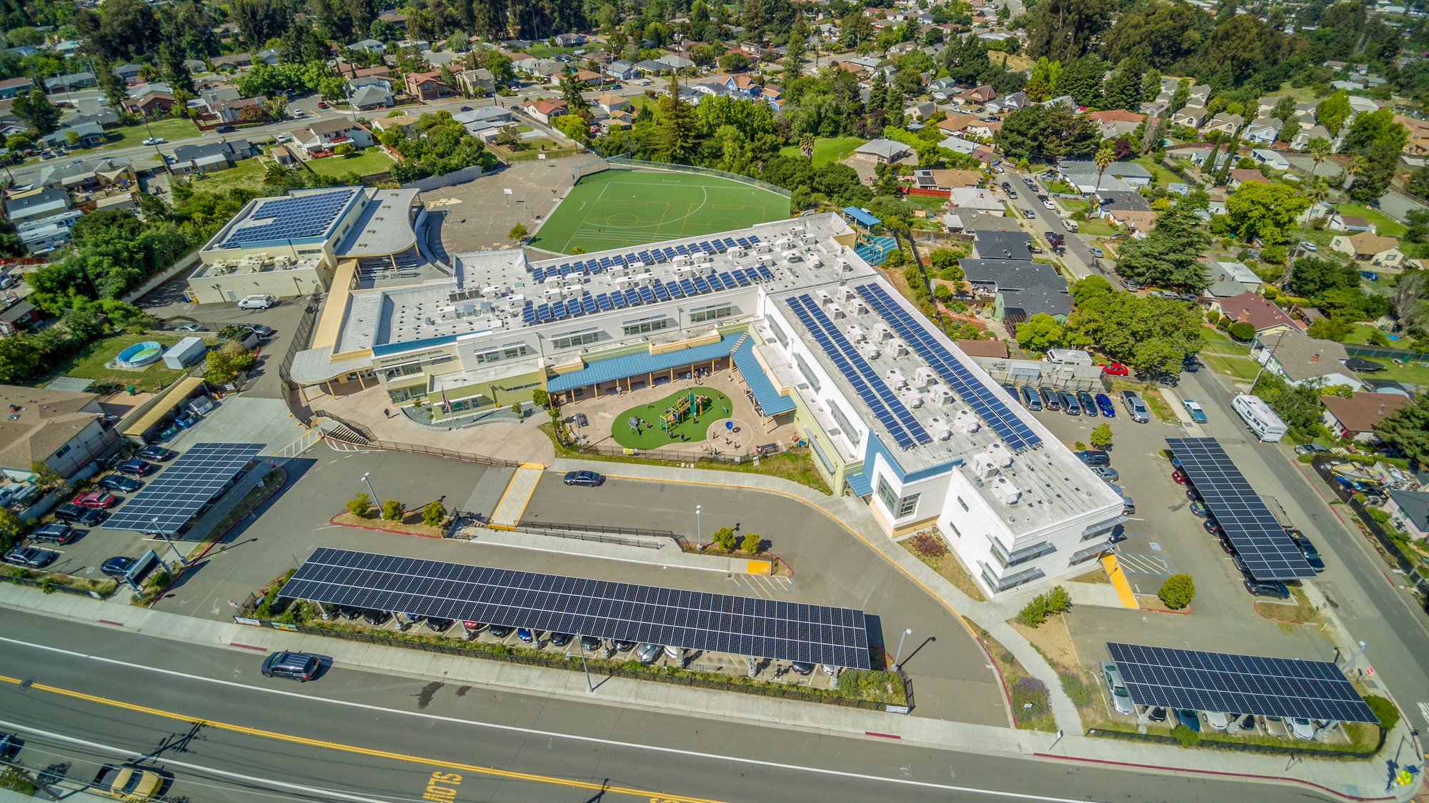 Aerial view of Hayward USD Fairview Elementary School showing campus buildings with rooftop and carport solar panels, playground areas, and surrounding neighborhood.