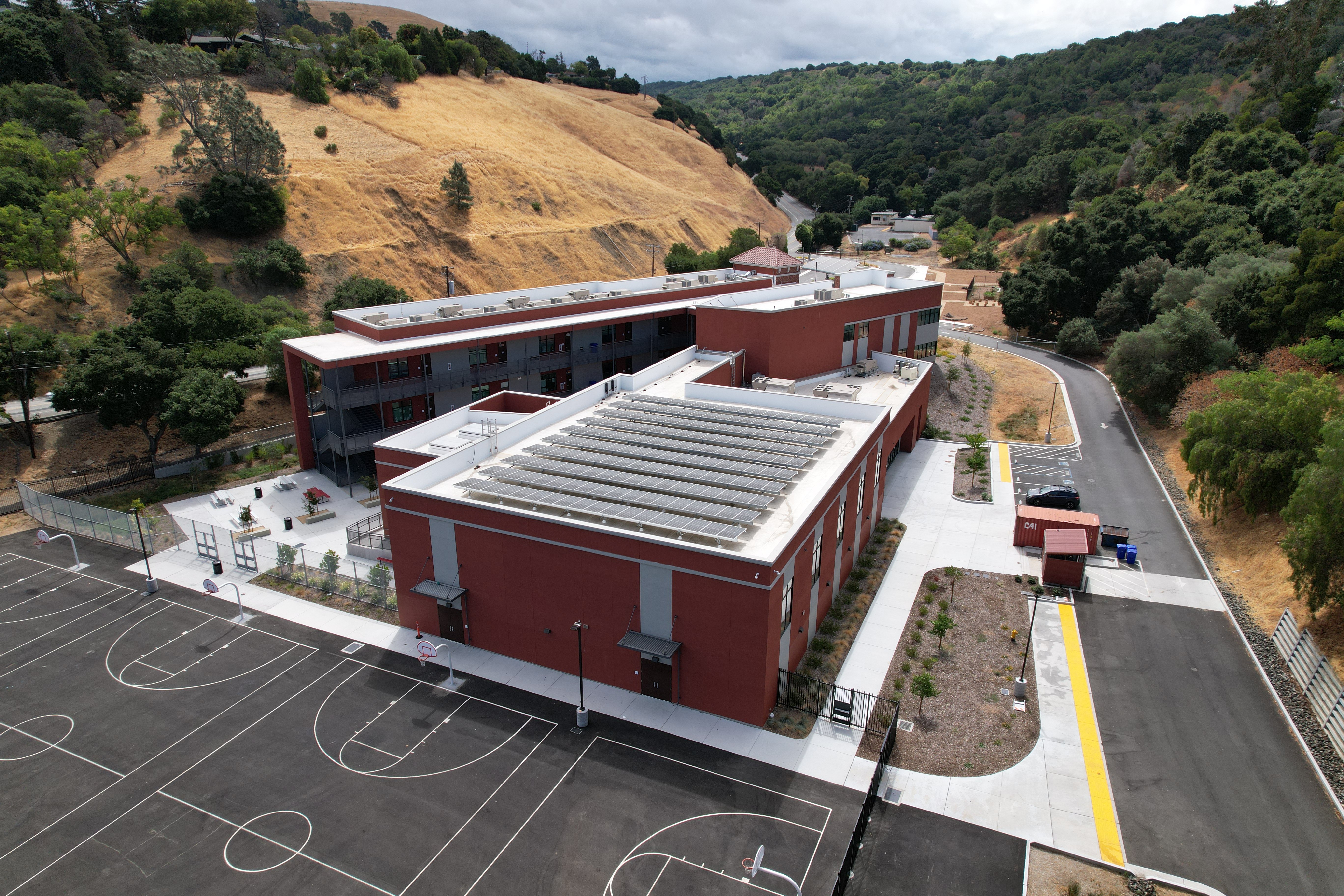 Aerial view of a modern red school building with rooftop solar panels and basketball courts, set against golden and green hillsides.