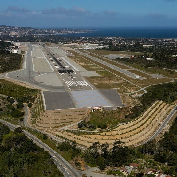 Aerial view of Monterey Regional Airport showing the main runway, taxiways, and surrounding terrain, with nearby hills, coastal landscape, and the Pacific Ocean visible in the distance.