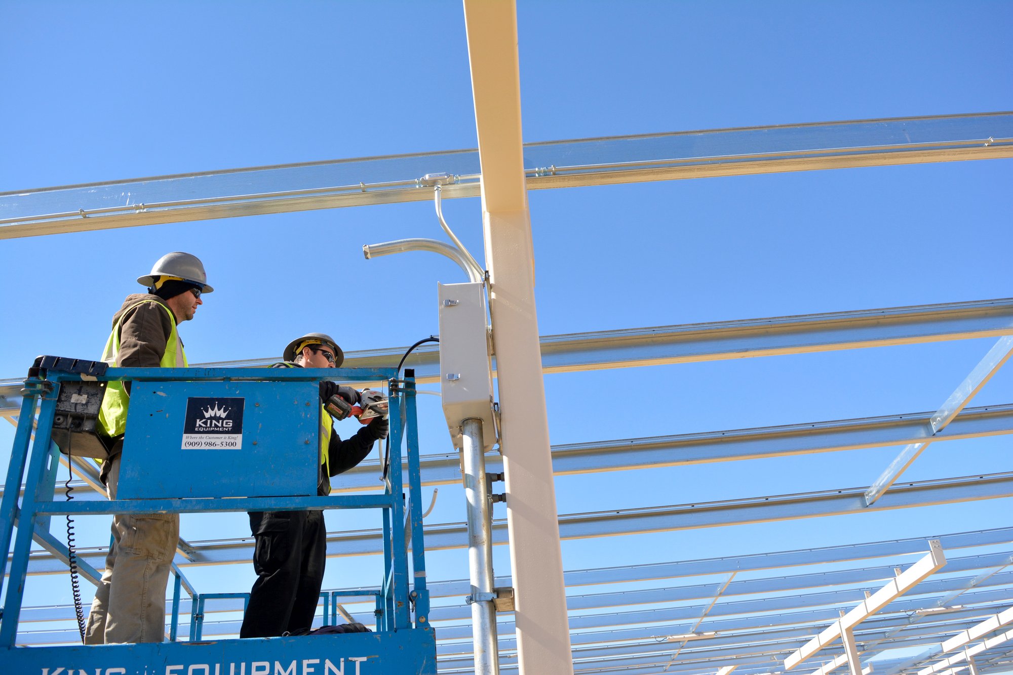 Workers installing electrical components on a solar carport structure at the Riverside County Ben Clark Training Center, using a lift to secure conduit and mounting hardware.