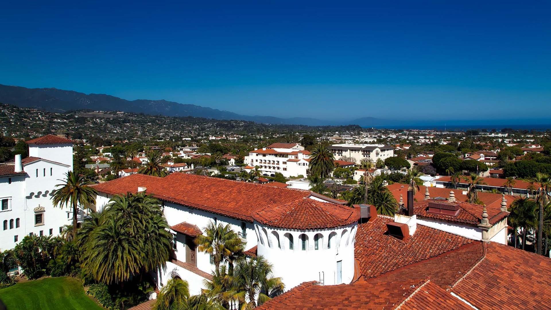Aerial view of Santa Barbara featuring red-tile rooftops, palm trees, and Spanish-style architecture with mountains and the Pacific Ocean in the distance.