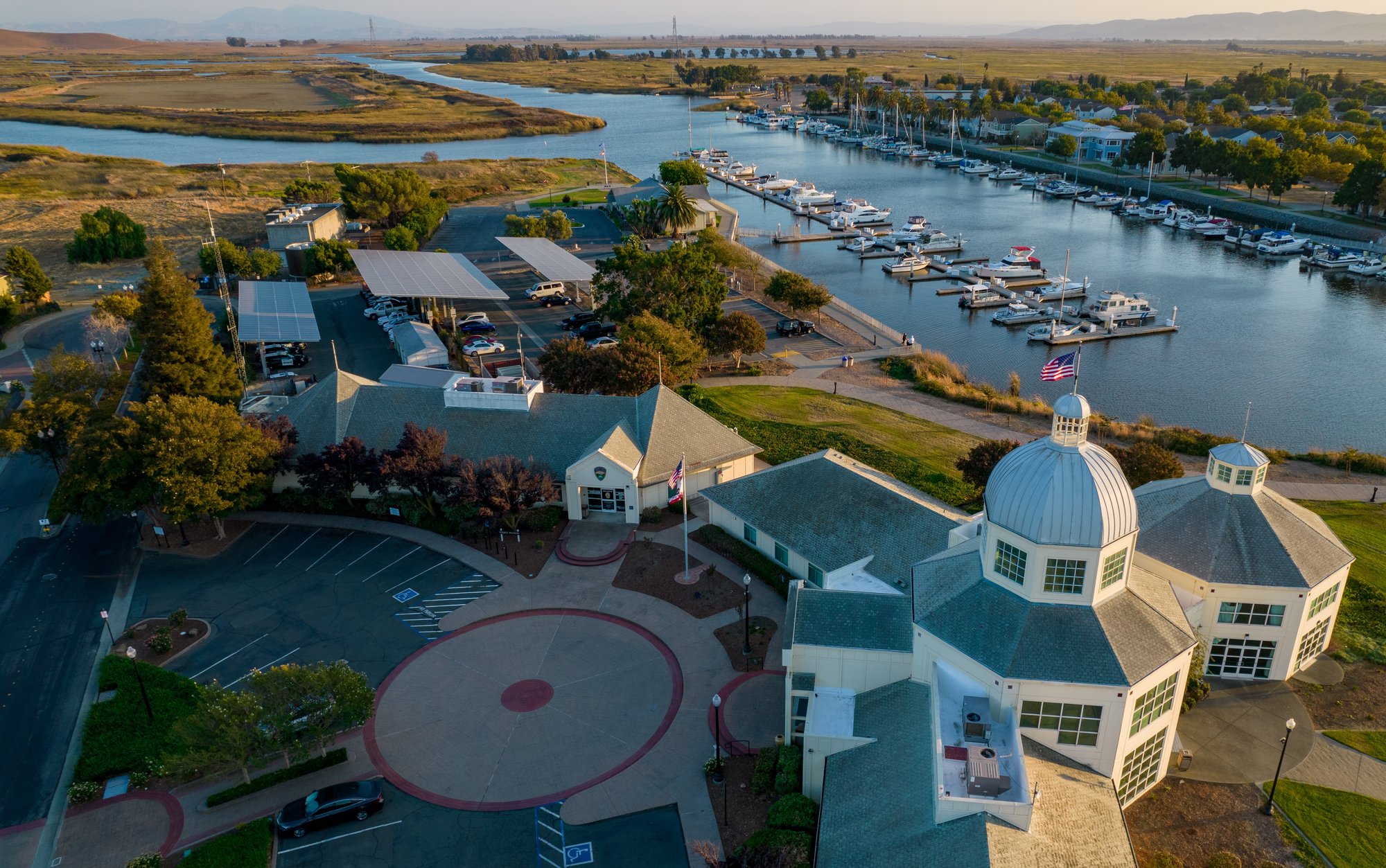 Aerial view of the City of Suisun waterfront featuring a marina with docked boats, a civic building with a domed roof, parking areas with solar carports, and surrounding wetlands at sunset.