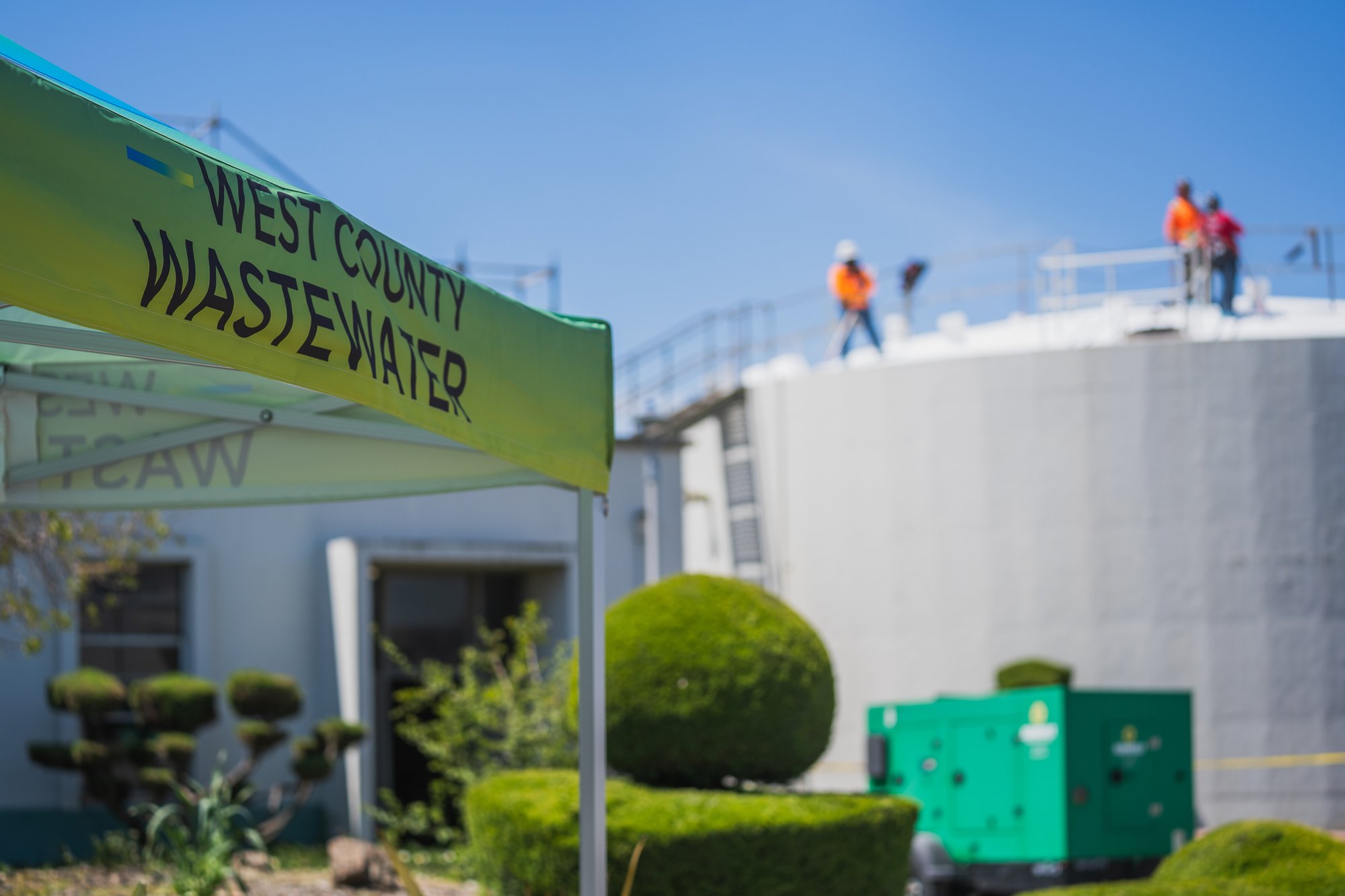 West County Wastewater facility with branded canopy in the foreground, maintenance staff working on an elevated tank, and backup generator equipment on site.