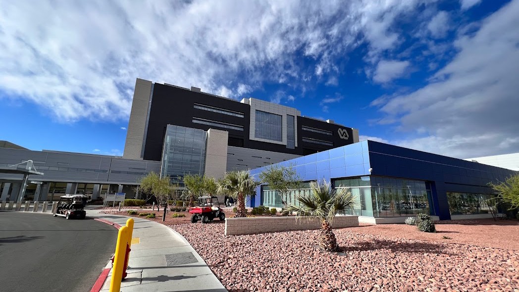 VA Southern Nevada Healthcare System hospital campus in Las Vegas with modern medical buildings, multiple American flags lining the entrance roadway, and desert landscaping under a clear blue sky.